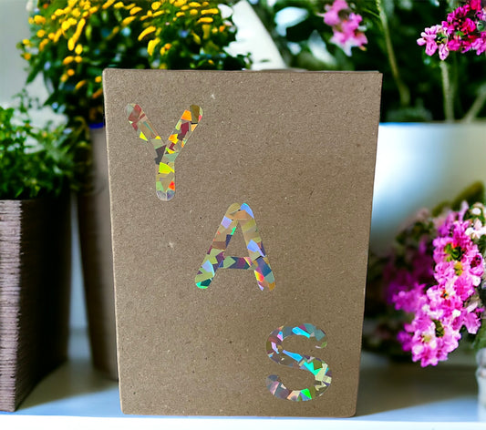 YAS HOLOGRAPHIC Greeting Card on a shelf full of colourful flowers