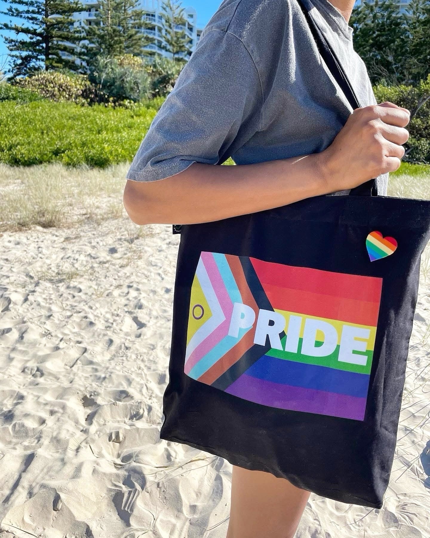 Person holding a black Pride tote bag with a rainbow heart pin on a progress pride flag design on a beach.