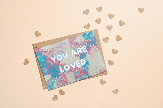 Greeting card with 'YOU ARE LOVED' text on trans pride flag coloured background with heart shapes.